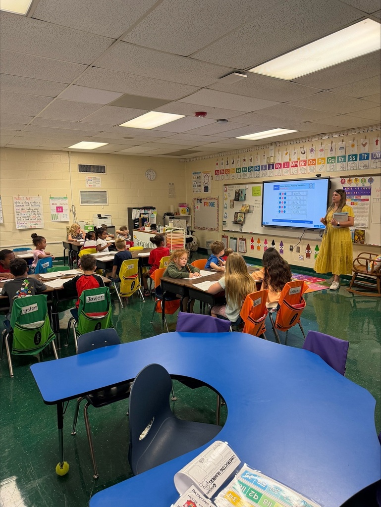 An indoor classroom with children seated at desks. A woman in a yellow dress stands near a projector screen.