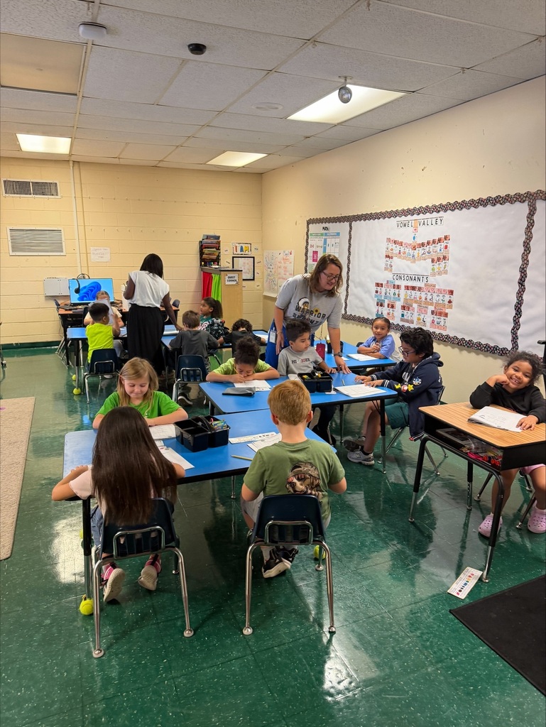 Kids sit at tables in a classroom, with an adult standing behind them, and a whiteboard in the background.