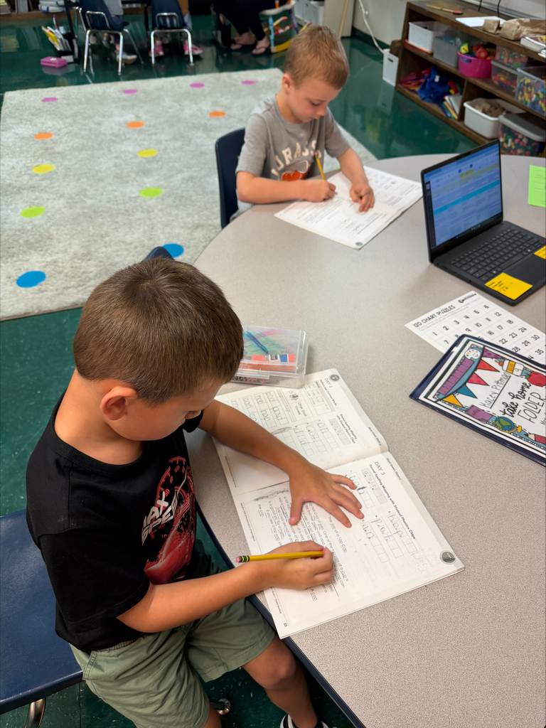 Two boys in a classroom, one using a laptop, the other writing on paper with a pencil.