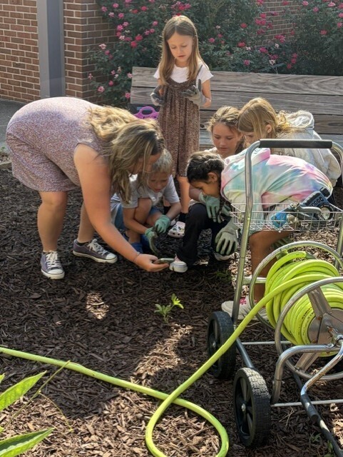 A woman and children plant a garden. A brick wall and greenery are in the background.