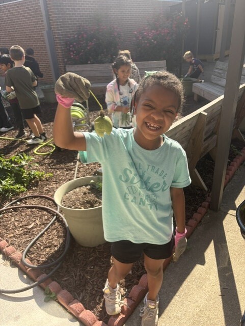 A young girl holds a green bean pod, smiling, in a garden with other children. Brick paths and wooden benches are nearby.
