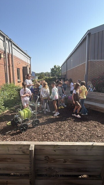 Several children and adults in an outdoor space with wooden fencing. A cart with a spool of green hose.