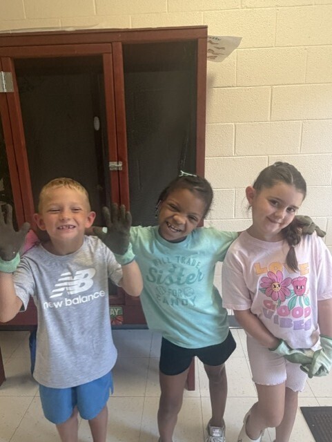 Three kids standing in a room with one holding gloves. Behind them, a wooden cabinet with glass doors.