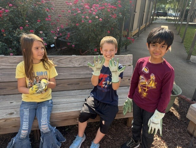 Three children in gloves sit on a bench in a garden. A girl and two boys pose for a photo.