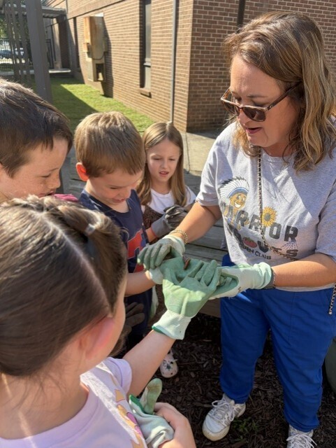 An adult woman helps several children, one holding a plant, put on gloves in a garden.