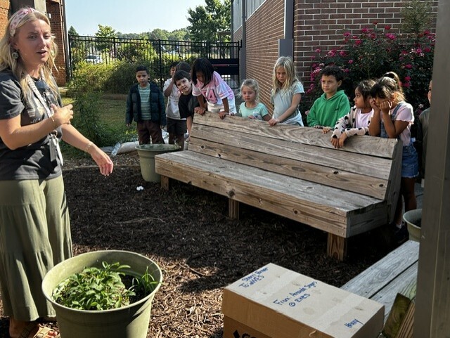 A woman speaks to a group of children standing behind a wooden bench. The woman holds a green bucket.