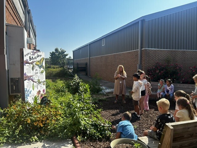 People gather outside a building, a woman stands near a mural while children sit and stand on benches.