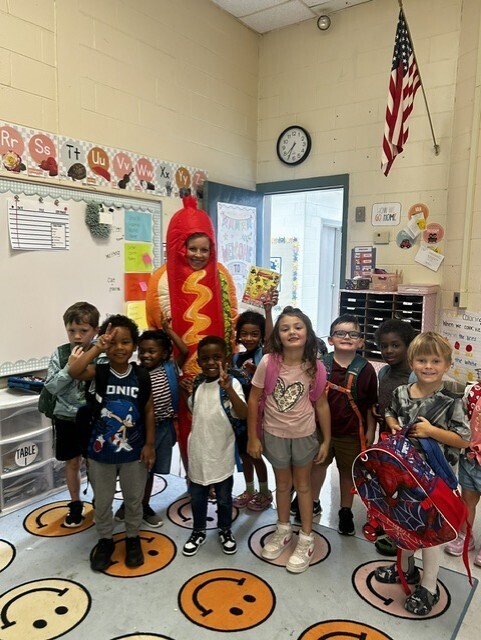 Group of children in a classroom with a mascot in front of a whiteboard and American flag.