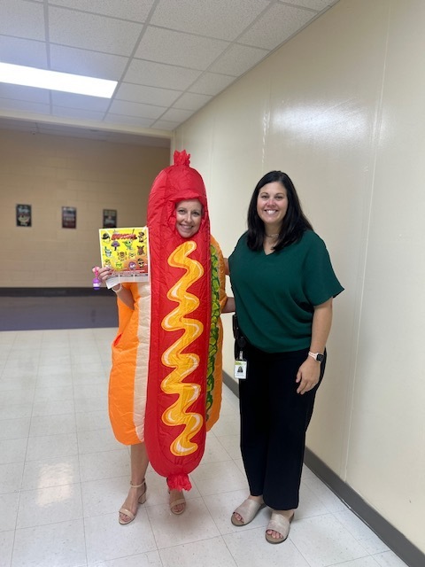 Two women, one in a hot dog costume and the other in a green shirt, stand in a hallway.