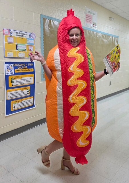 Woman in costume resembling a hot dog, standing in a hallway with posters on a white wall.