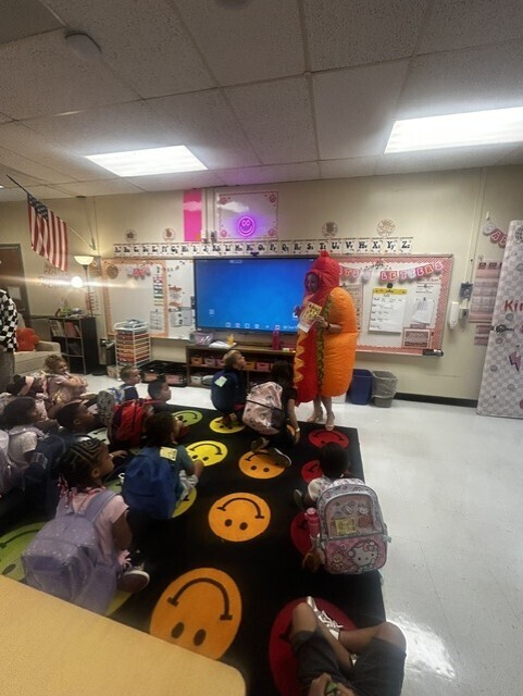 Classroom with children sitting on the floor watching a person in a costume. A projector screen is mounted on the wall.