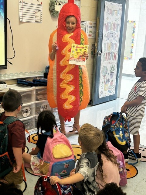 Woman in hot dog costume stands before children seated in circle. Children wear backpacks. Classroom has bulletin board and door.