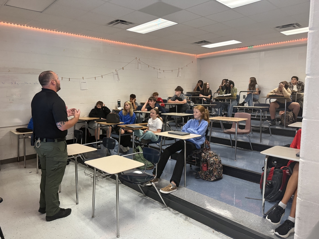 A man speaks to students in a classroom. Students sit on desks on raised platforms. White walls, white flooring, and ceiling lights.
