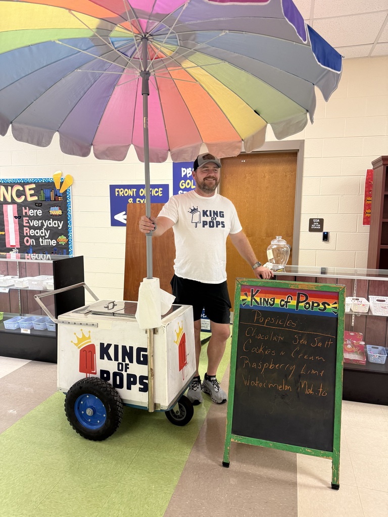 Person in a white t-shirt and shorts stands next to a cart with a rainbow umbrella.