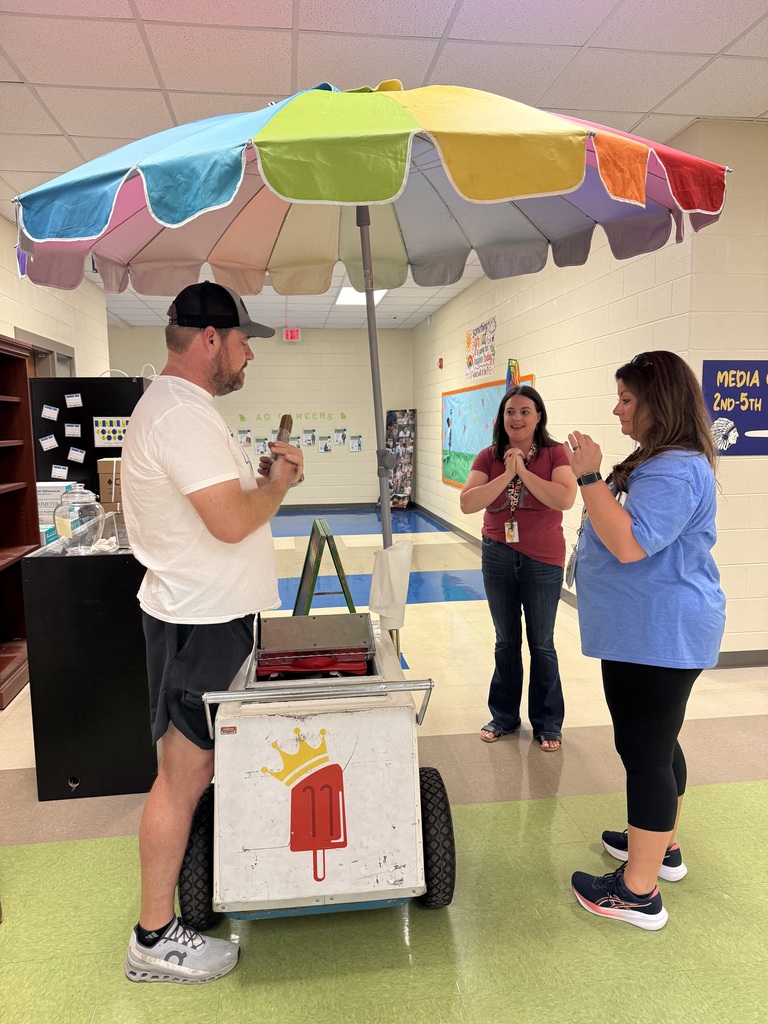 A man in a cap and sneakers stands at a colorful umbrella-topped ice cream cart, speaking to two women.