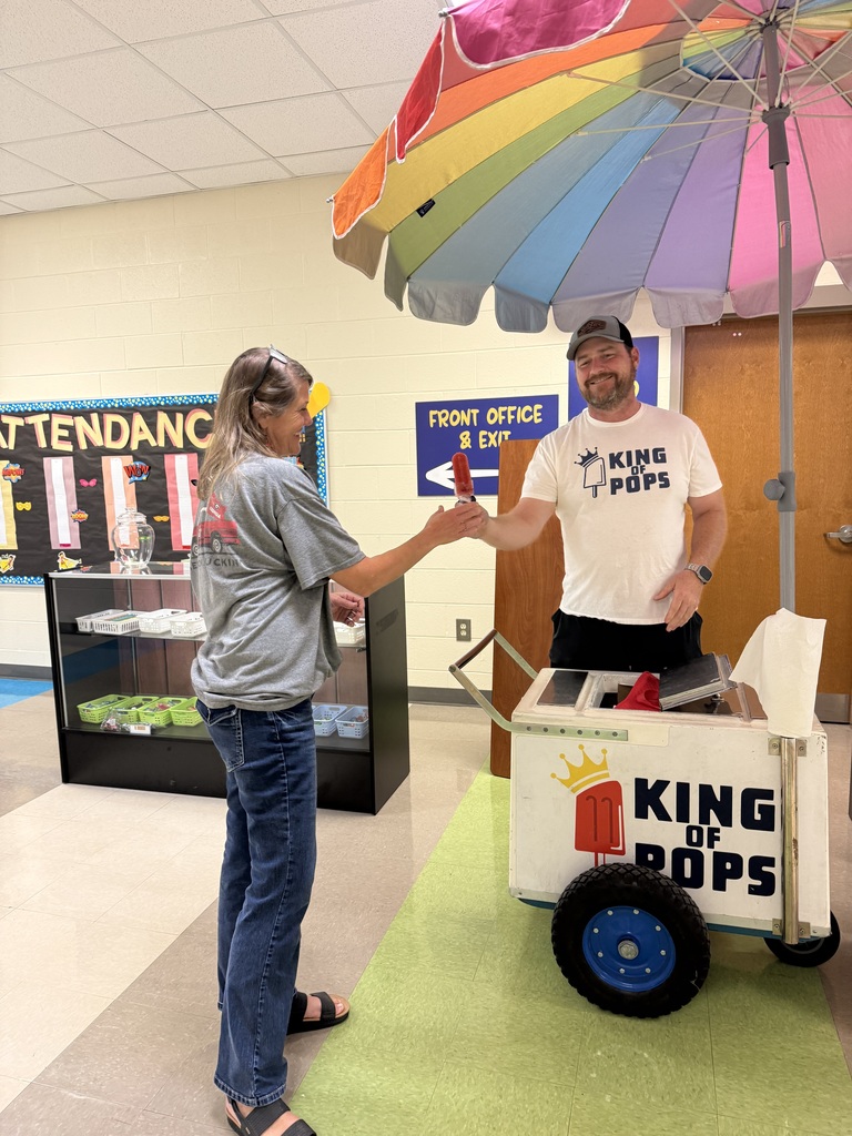 A man and a woman under a colorful umbrella, shaking hands. Behind them is a glass cabinet and a sign. The man operates a cart labeled "King of Pops."