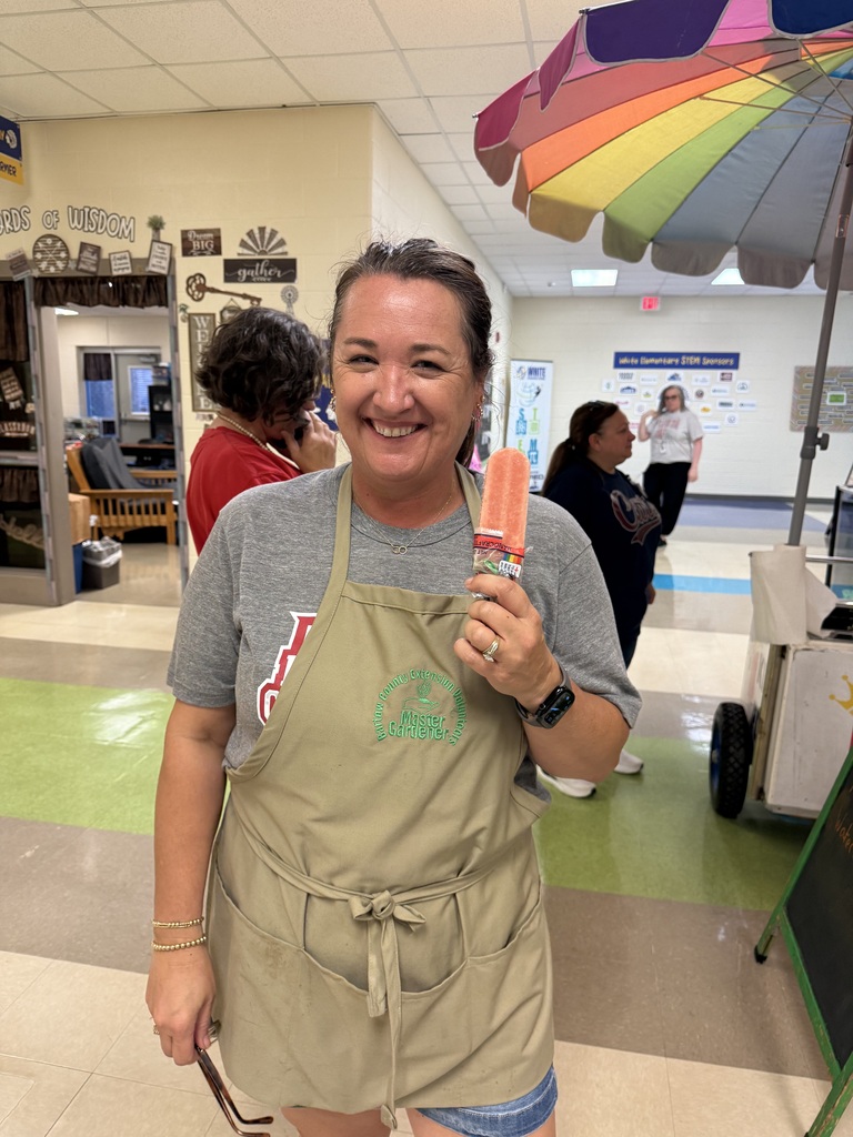 A woman in a khaki apron holds an ice pop. She smiles and stands next to a colorful umbrella.