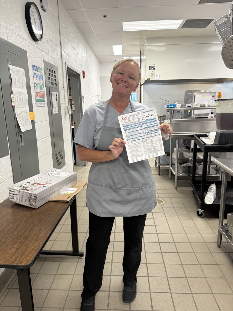 A person in a gray apron stands in a kitchen, holding a paper. Behind them, there is a table with boxes.