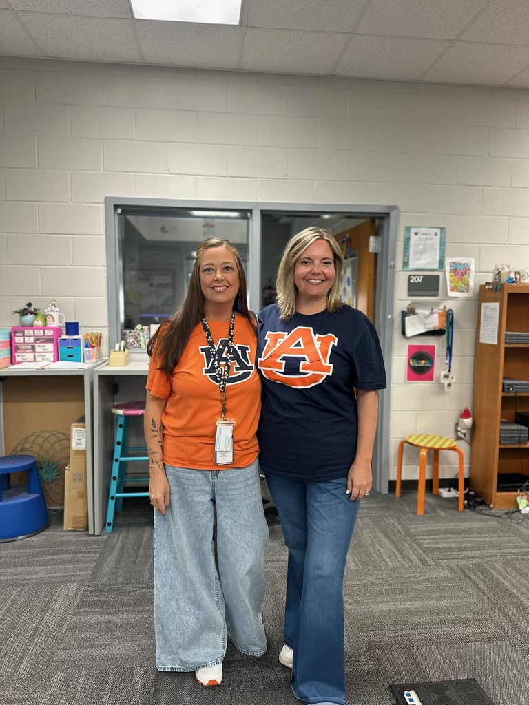 Two women stand in a classroom, one in an orange shirt and the other in a blue shirt.