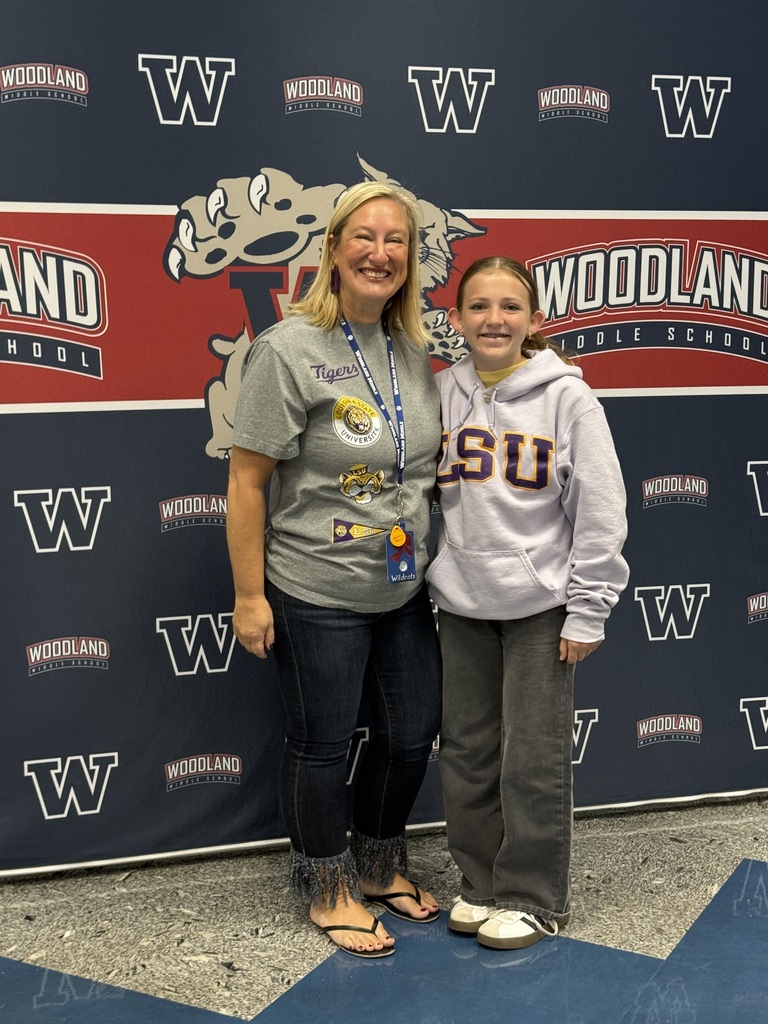 Two women standing in front of a Woodland backdrop, smiling. One wears a gray shirt, the other a gray hoodie.