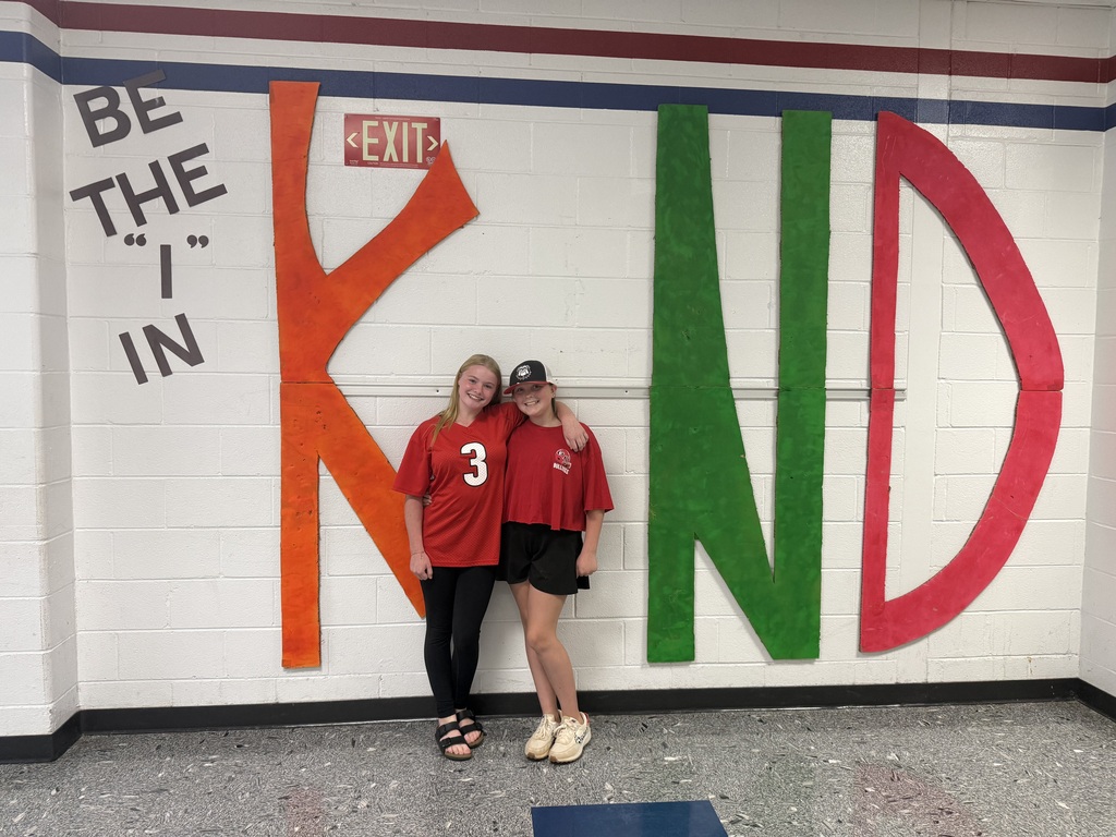 Two individuals in red shirts stand next to each other in front of a wall with large letters spelling out "KND".