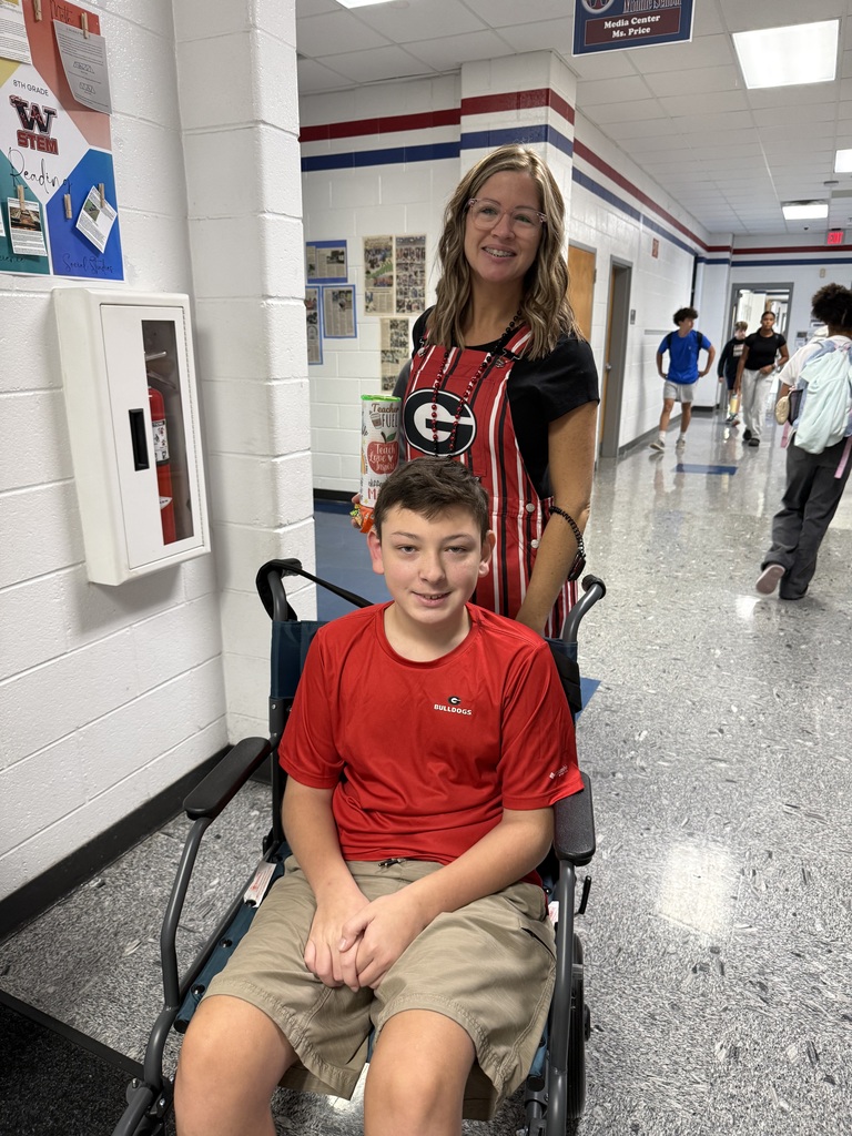 A young boy in a wheelchair and a woman wearing an apron stand in a hallway.