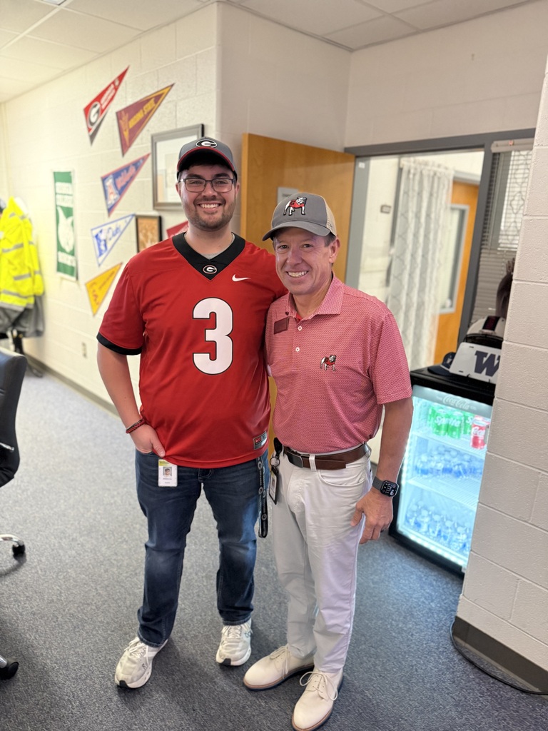 Two men stand next to each other in a hallway. One wears a red number 3 jersey, the other a pink shirt and hat.