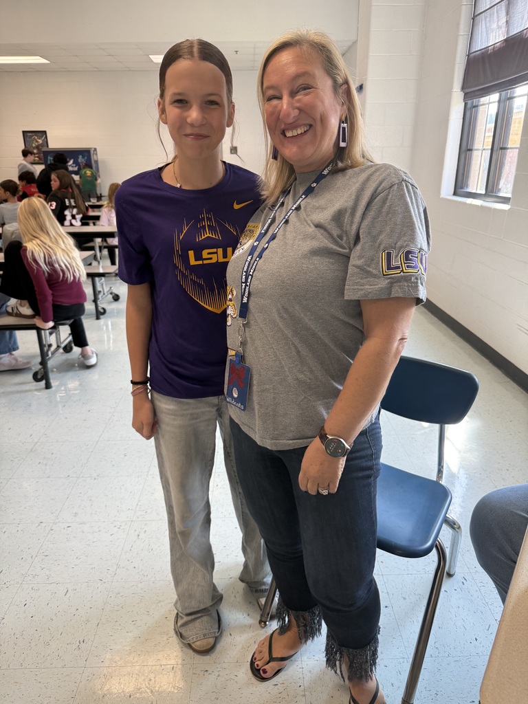 Two women in casual attire stand in a classroom, one wearing a purple shirt and the other a gray shirt. Behind them, people sit at tables and a blue chair is visible.