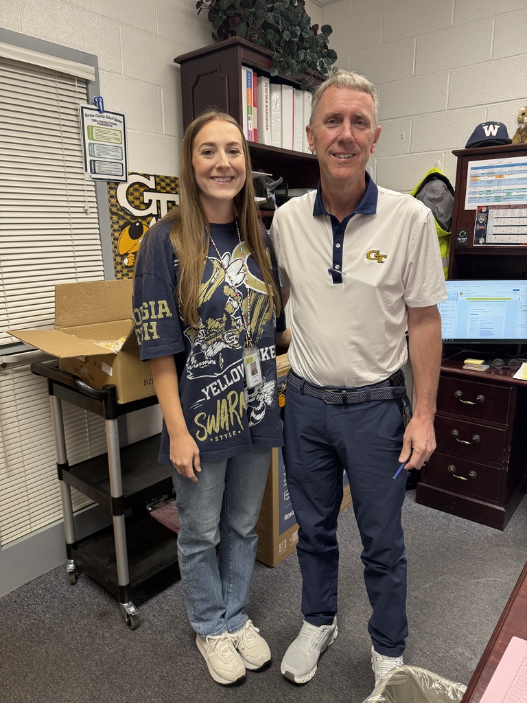 An adult man and a young woman stand together in an office, both wearing sneakers. A monitor, boxes, and a shelf with books are in the background.