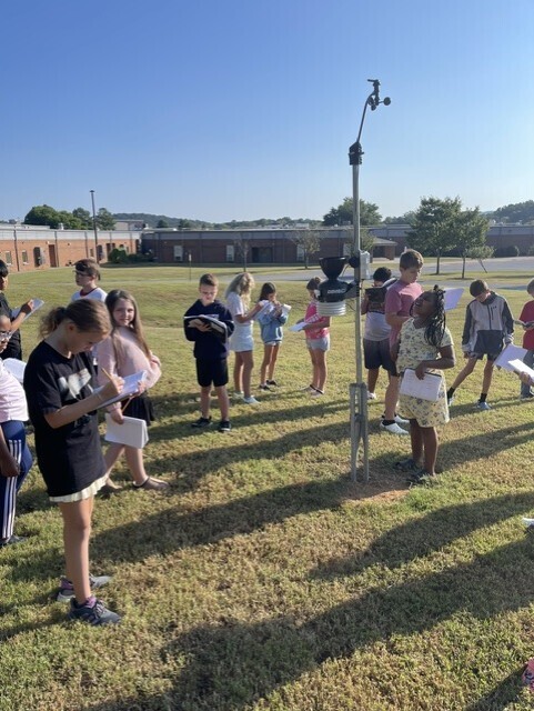 A group of children on a grassy field, holding papers and standing near a weather station.