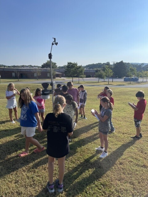 A group of children in various colored clothing gather around a tall device on a grassy field.