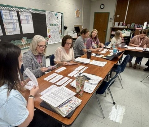 A group of adults in a room with a table, reading papers and books. Papers and books are on the table.