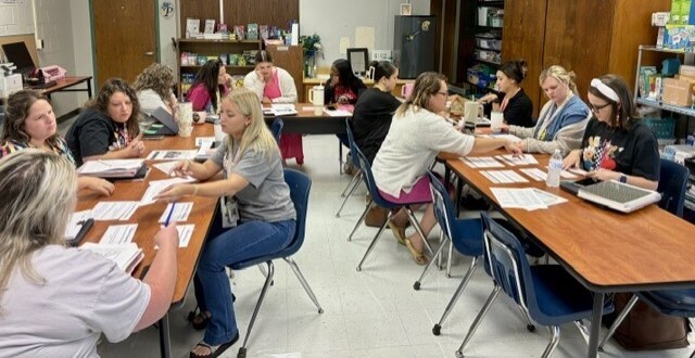 A group of women seated around tables in a classroom. Some are writing while others hold papers.