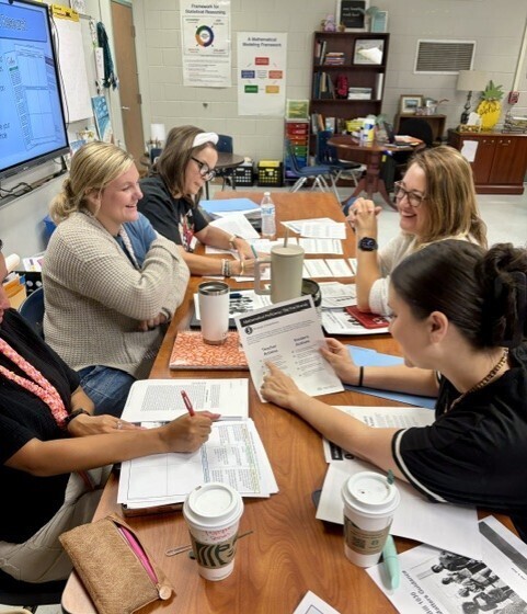 Five women sit around a table, reviewing papers. One holds a pen. Two cups, some papers, and a handbag are on the table.