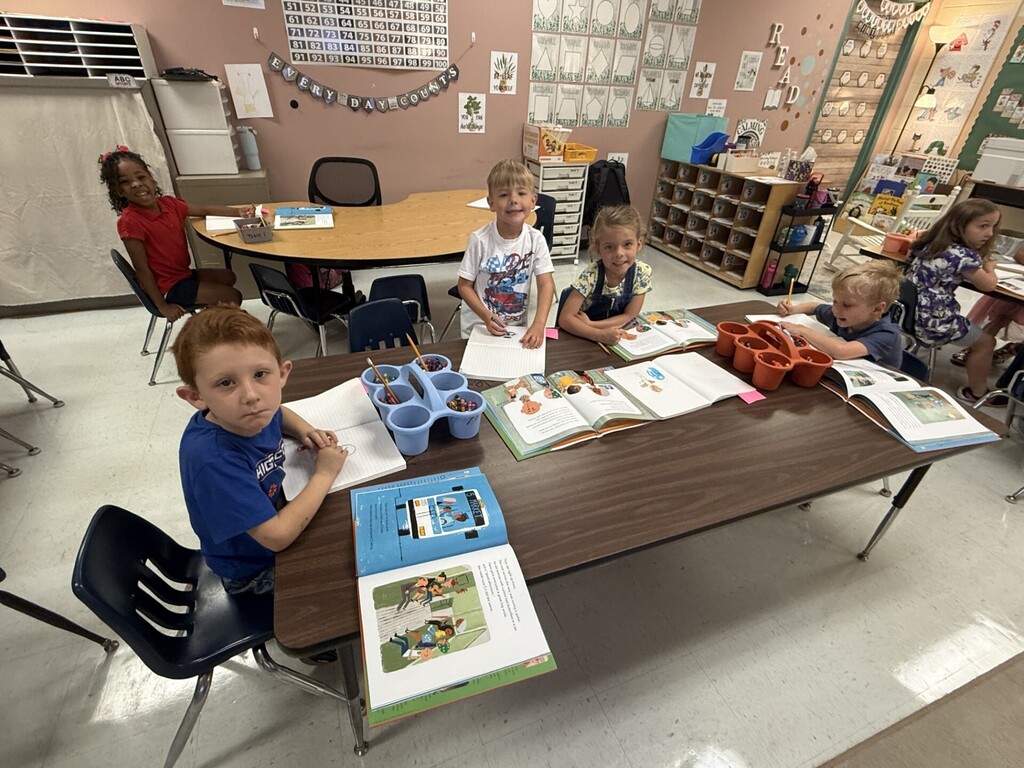 Several children seated at desks, working with books, crayons, and paper. Pink wall with decorations.