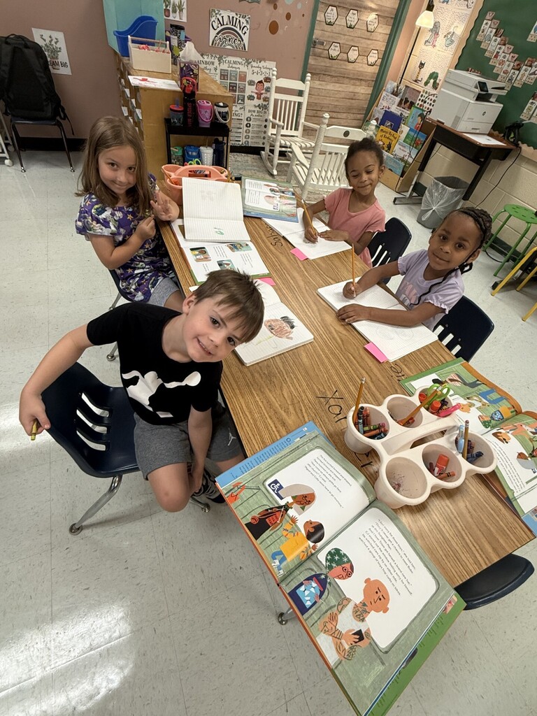 Four children sitting at a long desk in a classroom. A boy wearing a black t-shirt sits on the left, with three girls on the right.