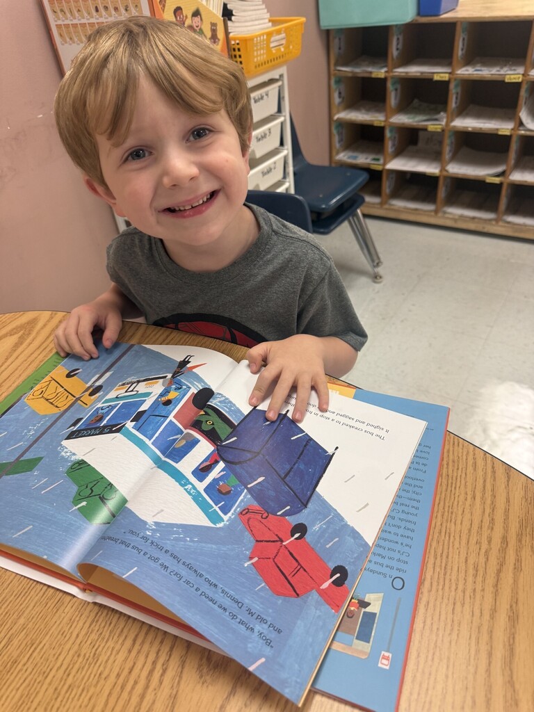 A child smiles while sitting at a table, reading a colorful book with illustrations of vehicles.