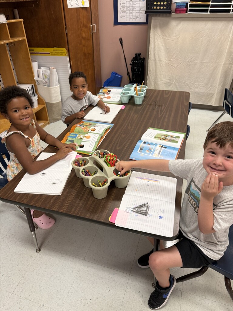 Three children at a table with open books and pencils in a classroom. One child is smiling.