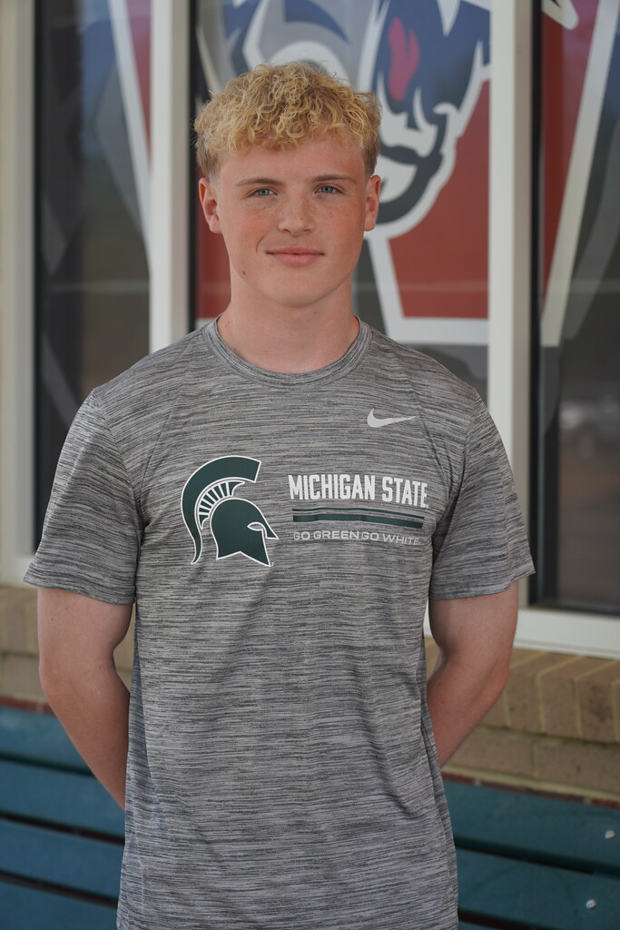 Man with blonde hair wearing a gray Michigan State shirt stands with hands on hips. Behind him, a window displays a Michigan State logo.