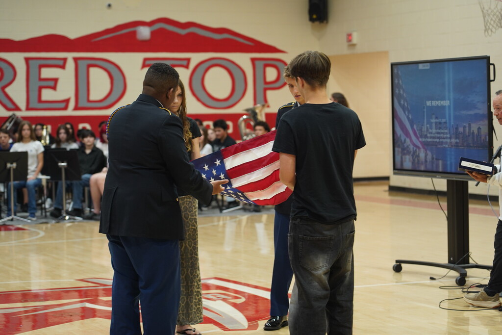 People stand in a gymnasium. A man hands an American flag to another man. A large screen is on the wall.