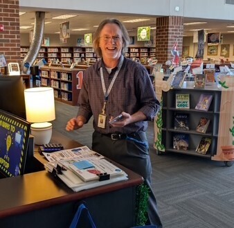 A person stands at a desk with a computer, lamp, and books. Shelves with books are in the background.