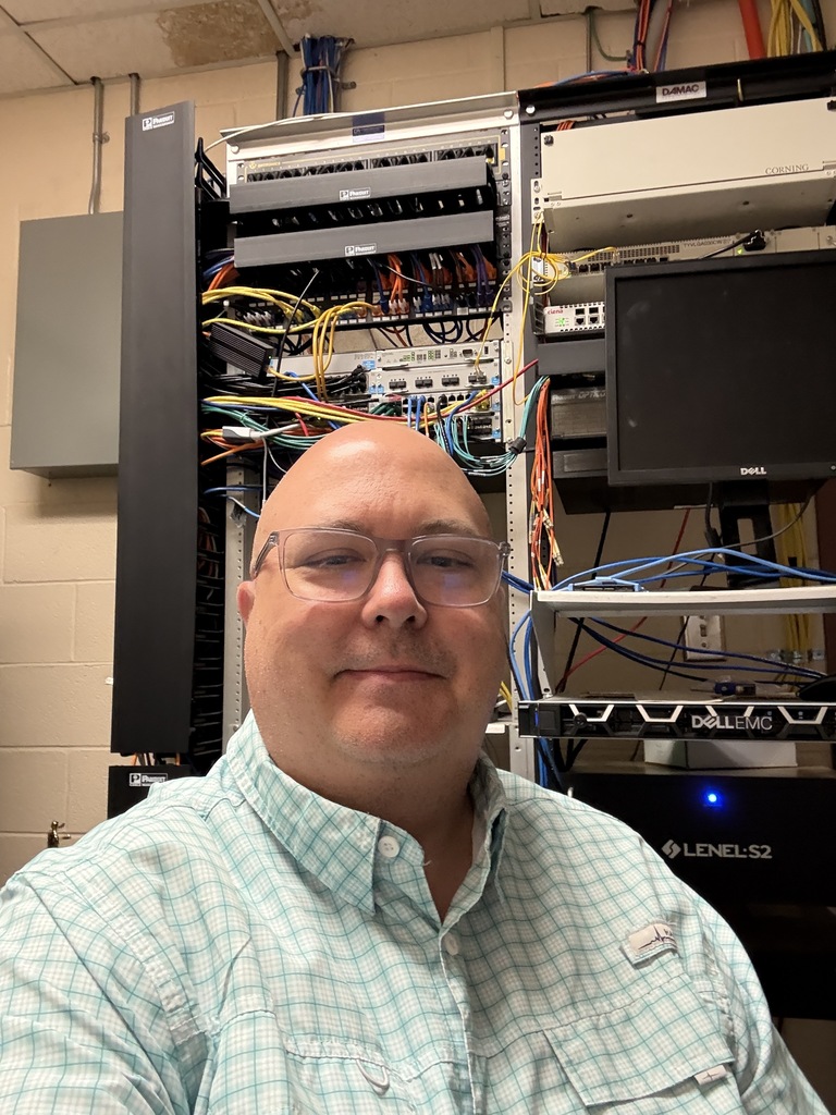A smiling man wearing glasses stands in front of a server rack with wires and a monitor.