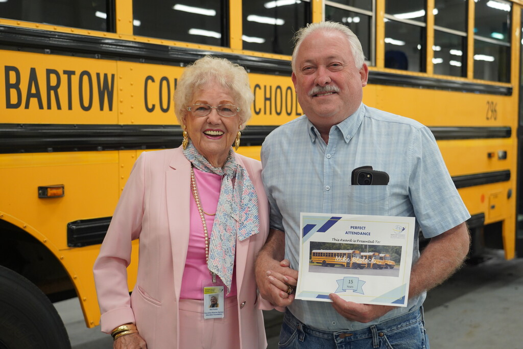 A woman and a man standing next to a school bus holding a certificate.