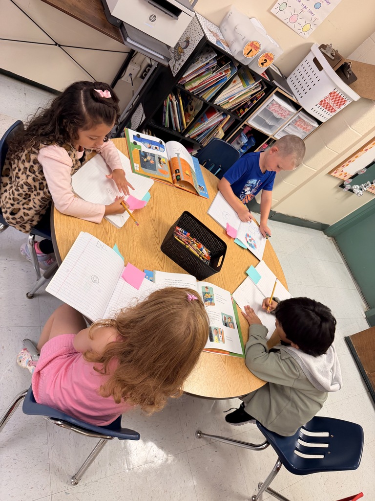 Four young children sit around a round table, working on school assignments, with books, papers, and pens.