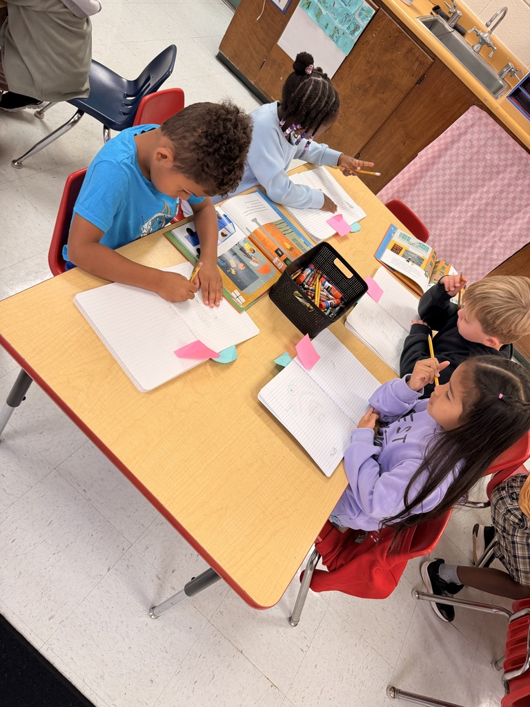 Four children seated around a table in a classroom. Each child is engaged in writing or drawing on paper.