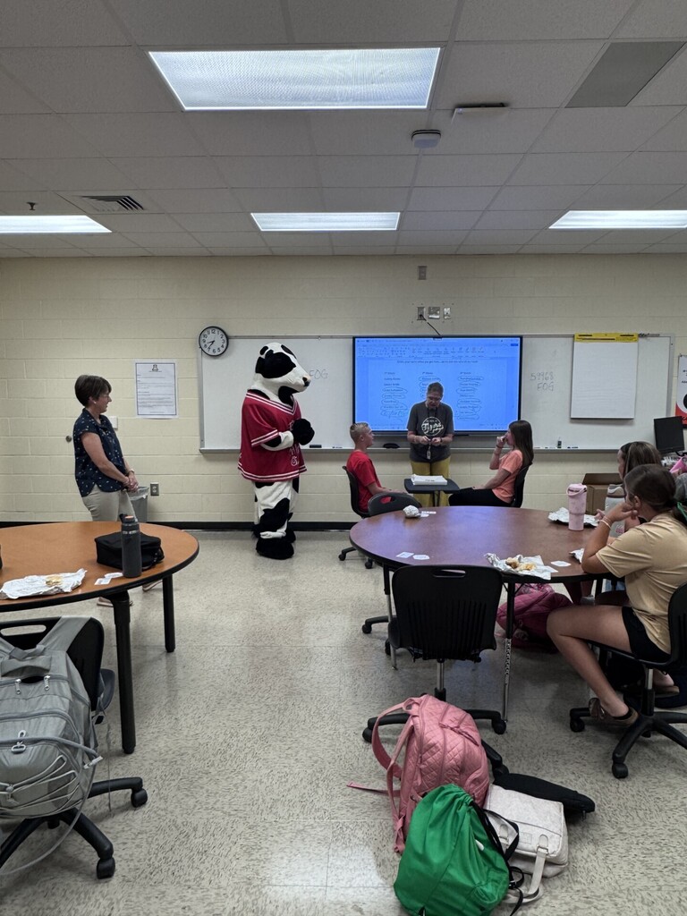A classroom with tables and chairs, a mascot, and a projector screen on the wall.
