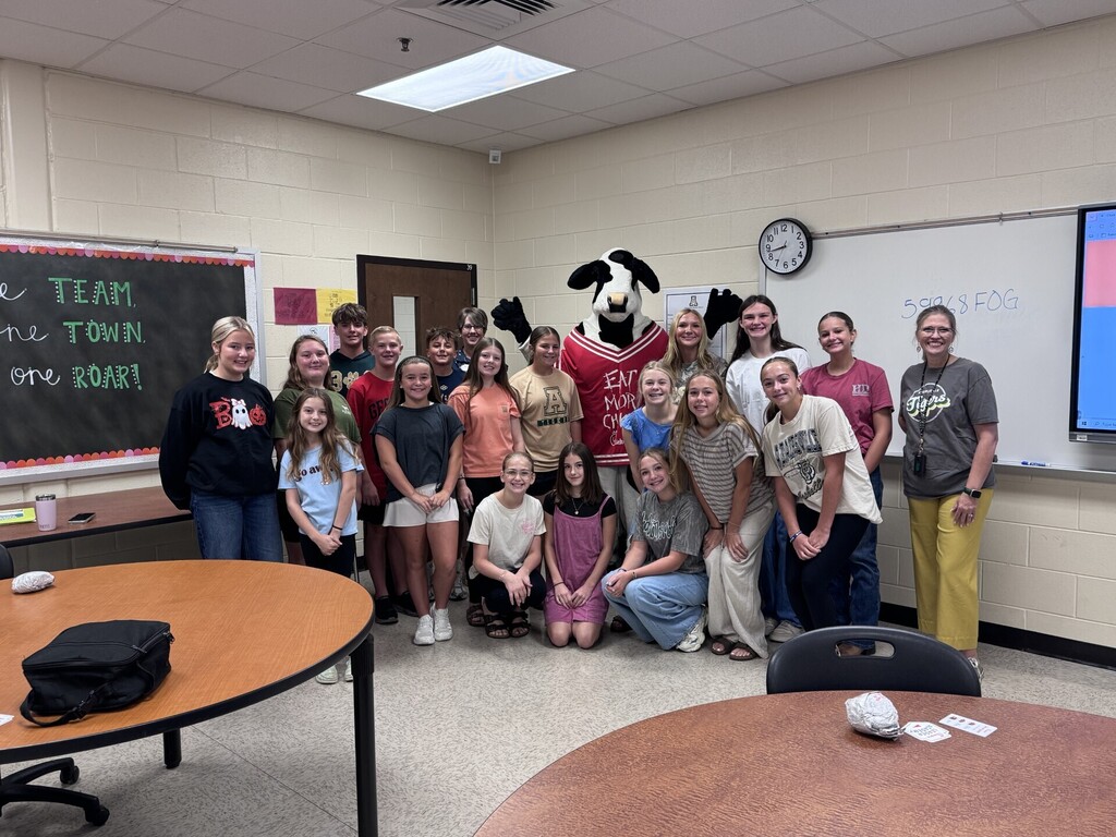 Group of young people with a mascot, in a classroom with desks, a blackboard, and a whiteboard.