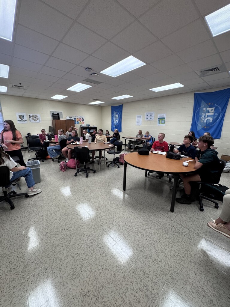 An indoor classroom with round tables, chairs, and several people seated and standing. A white floor, ceiling lights, and banners hang from the walls.