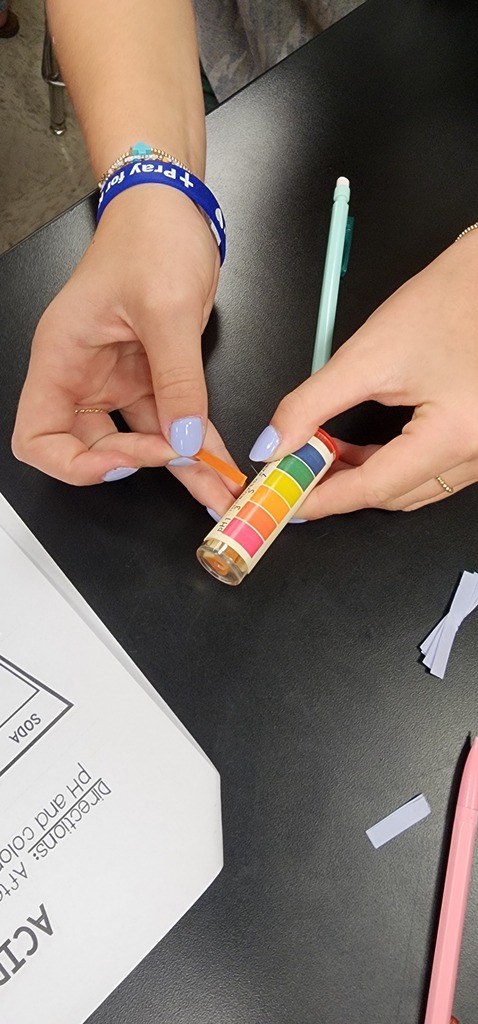 A person with blue nails holds a small, multicolored stick over a white sheet of paper. Hands next to pens and cut paper.