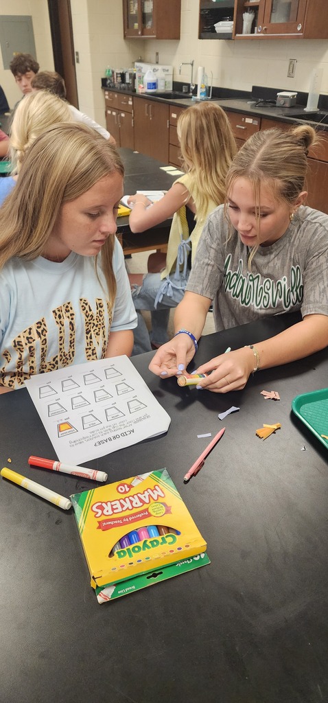 Two girls at a table with art supplies. One holds a box of colored pencils. Background: a classroom.